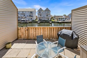 Une terrasse avec une table et des chaises en verre, un barbecue abrité et une vue sur une marina, avec des bateaux et des bâtiments en arrière-plan.