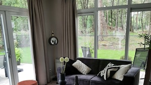 Un salon moderne avec un canapé, une table basse ronde et un pouf. De grandes fenêtres offrent une vue sur un jardin.