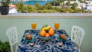 A table with a floral tablecloth, two white chairs, a bowl of fruit, two glasses of orange juice, and two small glasses of a dark beverage, overlooking a mountainous landscape with a bridge.