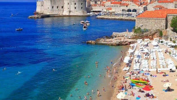 A coastal city with a historic fortification, a bustling beach with sun loungers and umbrellas, and boats in the water.