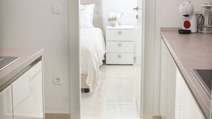 Quarto moderno e minimalista com cama, mesa de cabeceira e criado-mudo. Uma porta branca dá acesso ao banheiro, que possui vaso sanitário e pia. O quarto tem um espelho grande e um relógio na parede.