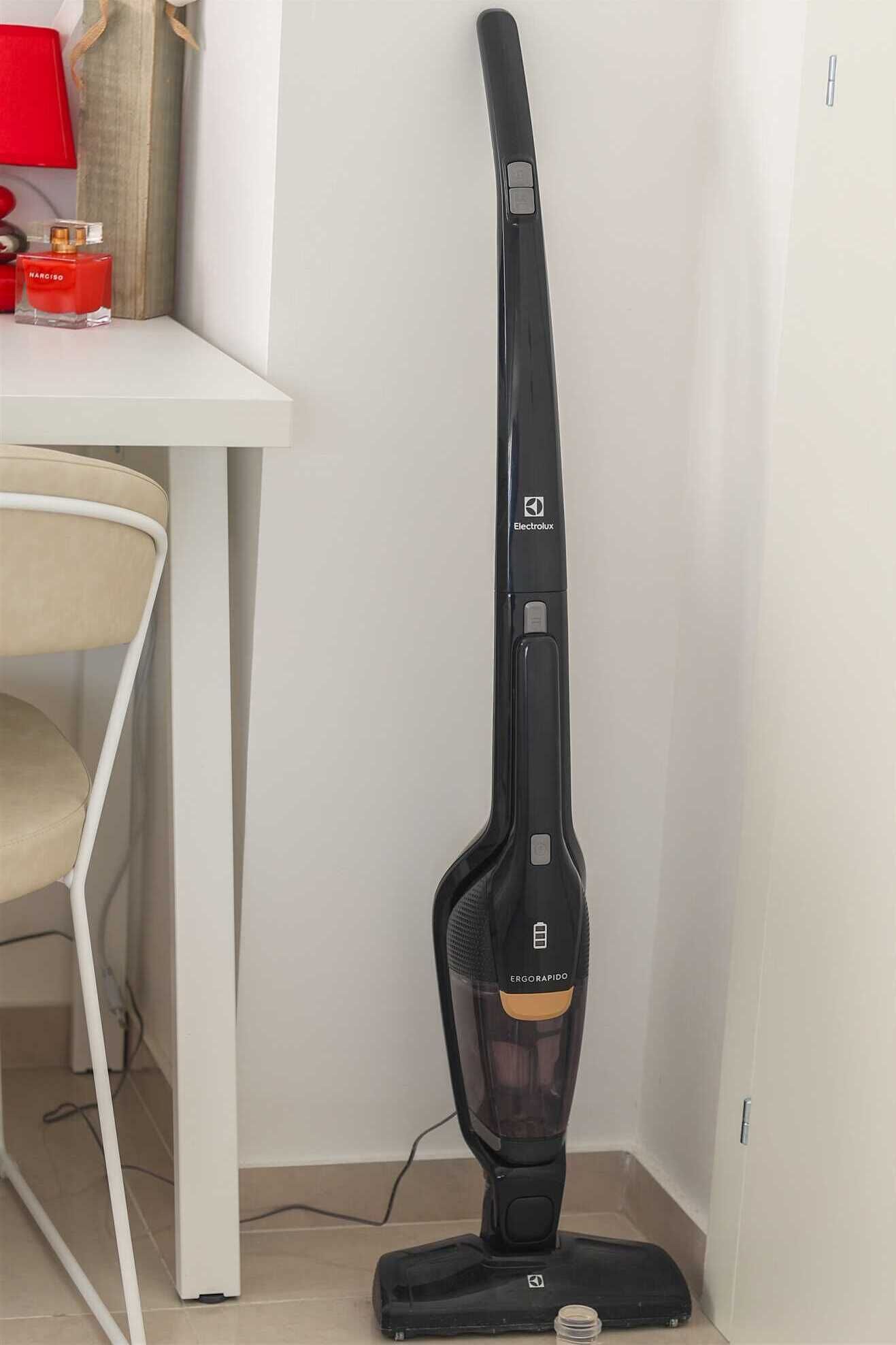 A black vacuum cleaner with a transparent dust container, placed in a corner of a room with a white table and a beige chair.