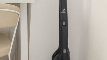 A black vacuum cleaner with a transparent dust container, placed in a corner of a room with a white table and a beige chair.