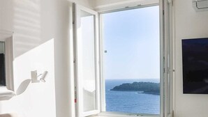 Quarto com uma janela grande, uma cama, uma cadeira, uma escrivaninha e uma televisão.