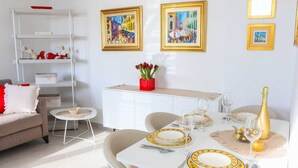 Uma sala de jantar com uma mesa branca posta para quatro pessoas, com pratos amarelos e dourados, uma garrafa de champanhe e um vaso de flores vermelhas.
