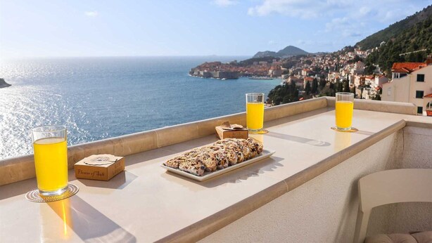 Uma varanda com vista para o mar e uma cidade, com uma bandeja de comida e dois copos de suco de laranja.