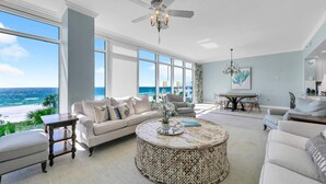 Een ruime woonkamer met een groot raam met uitzicht op een strand, een plafondventilator en een ronde salontafel met een decoratief middelpunt.