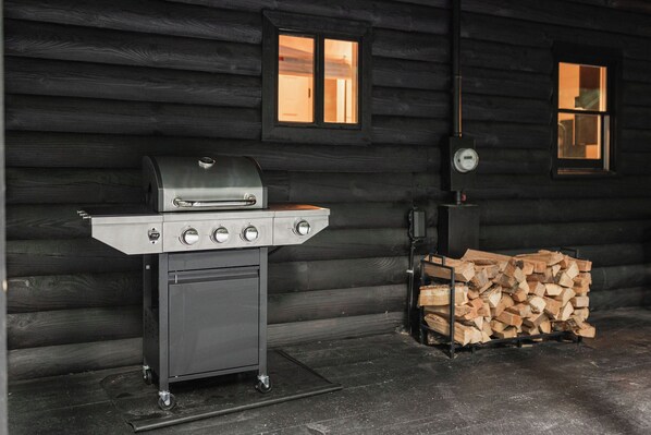 Una parrilla portátil con cuatro quemadores y un horno integrado, sobre un carrito con ruedas, al lado de un montón de leña, frente a una cabaña de troncos con dos ventanas.