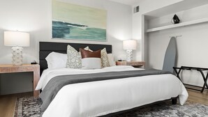 Une chambre d’hôtel moderne avec un grand lit, deux tables de chevet, une petite table, une planche de surf accrochée au mur et un tableau représentant une scène de plage.