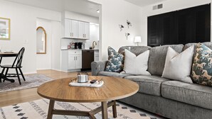 Un salon avec un canapé gris, une table basse en bois et un tapis à motifs. On aperçoit une cuisine à l’arrière-plan, avec des meubles blancs et un plan de travail noir.