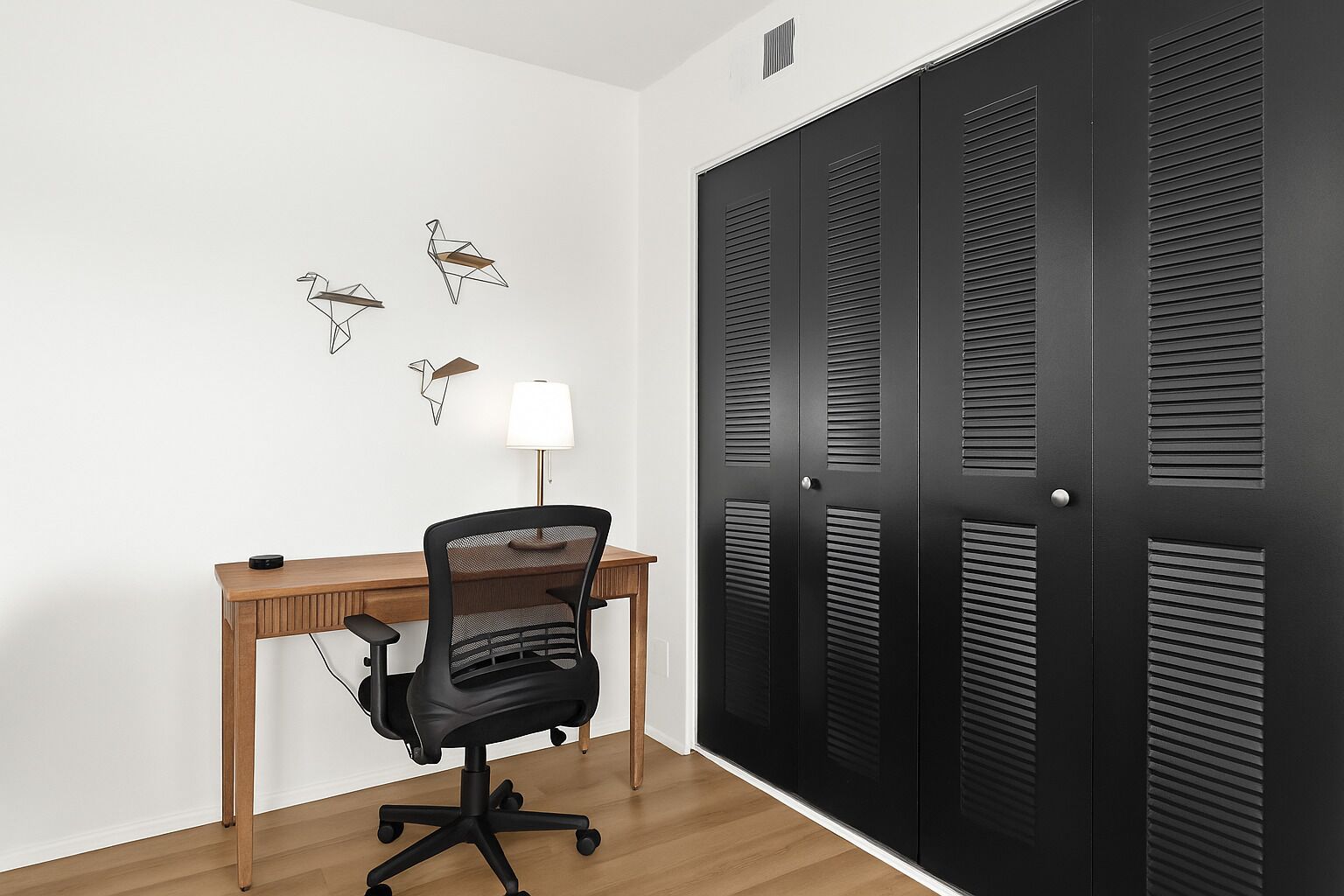 A room with a wooden desk, a black office chair, a white lamp, and a set of black louvered doors.