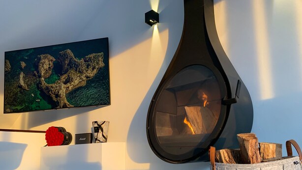 Un soggiorno moderno con camino, televisione a parete che mostra una scena costiera e un cesto decorativo per legna.