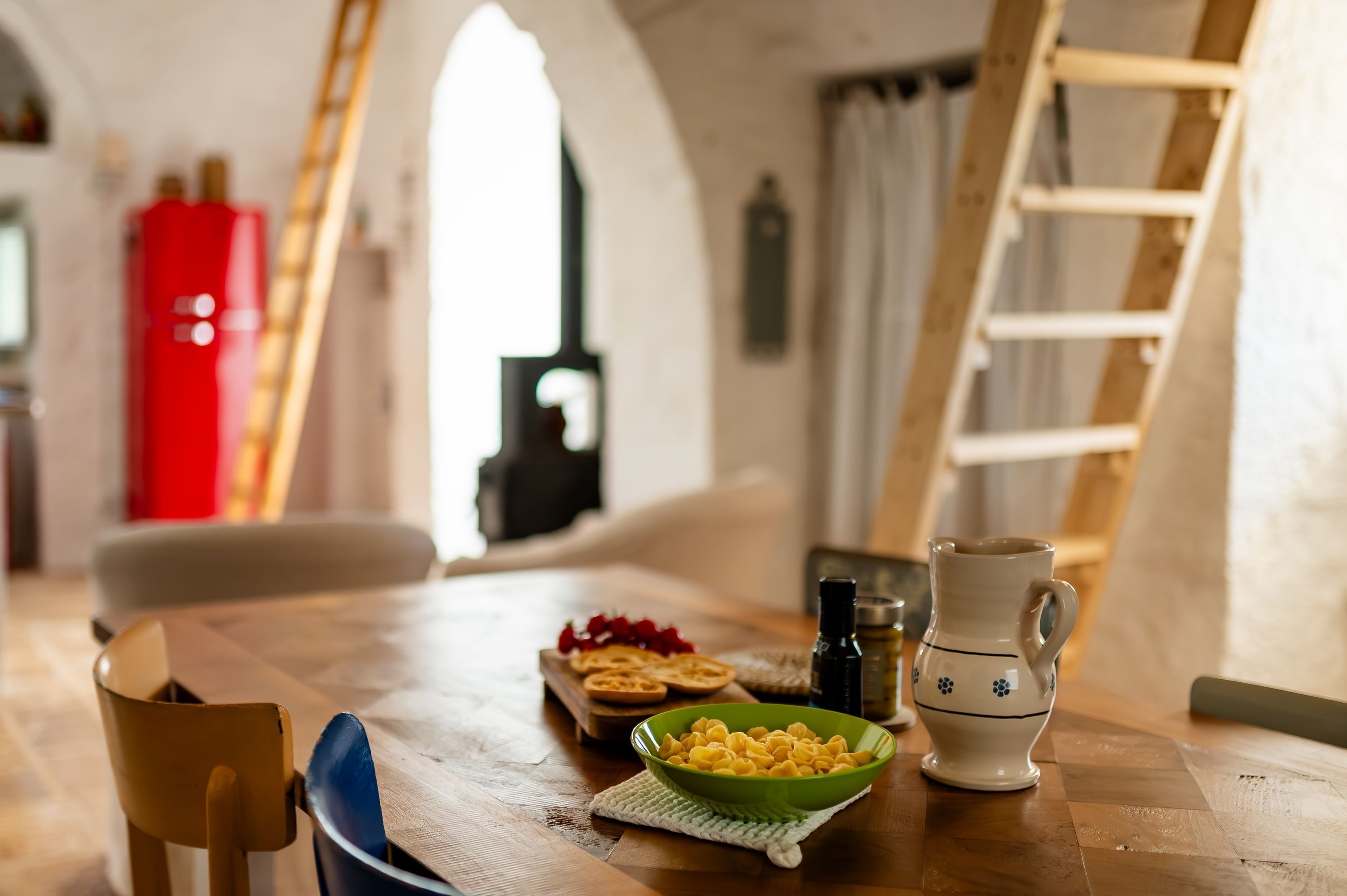 A wooden table with a bowl of pasta, a white pitcher, and a red appliance in the background.