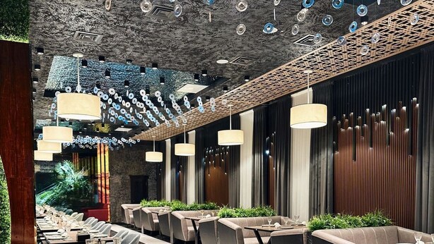 Een modern restaurant met lange gedeelde tafels, individuele zitplaatsen en een bijzonder plafond met hangende elementen.