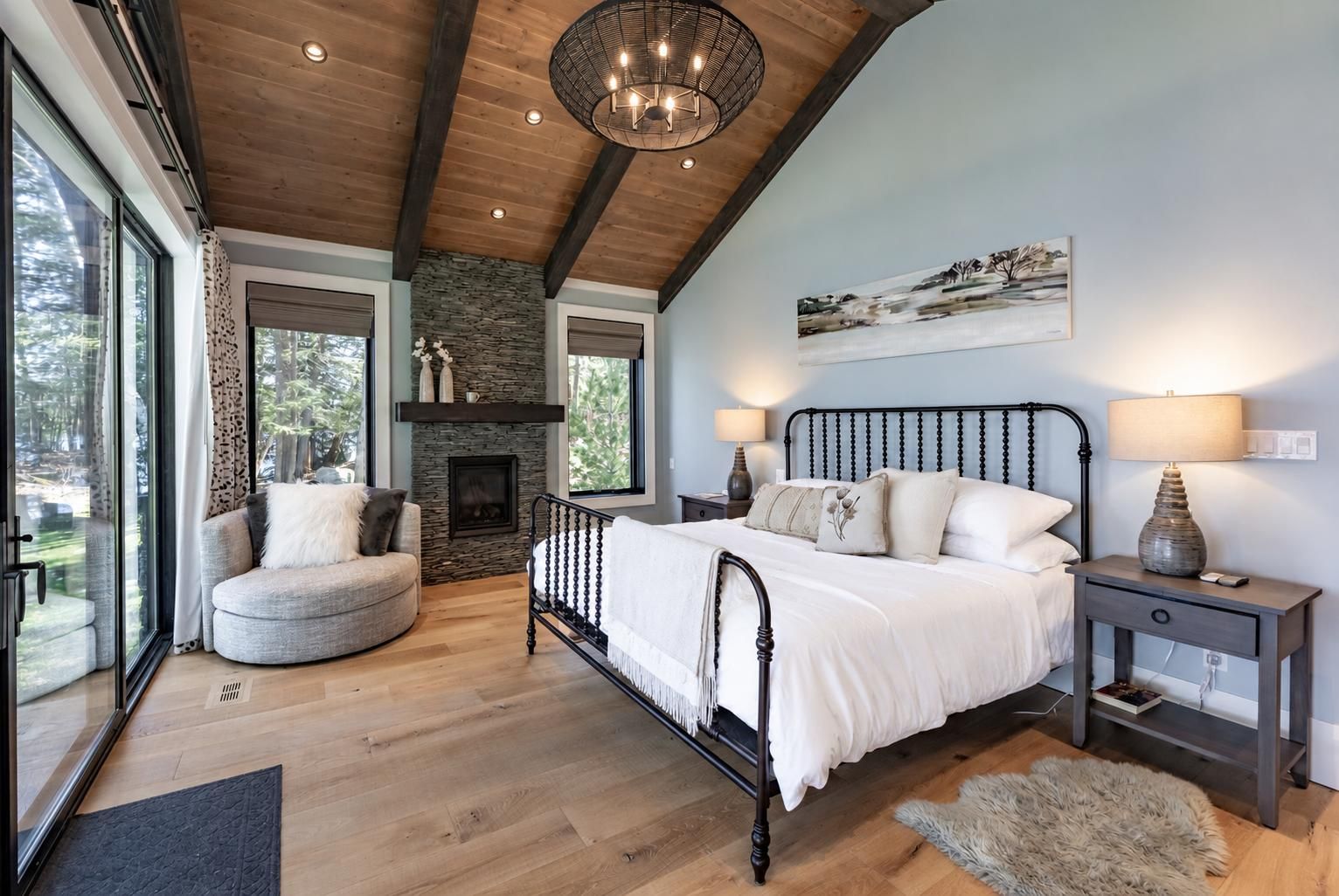A bedroom with a stone fireplace, a bed with white bedding, a nightstand with a lamp, and a view of the outdoors through large windows.