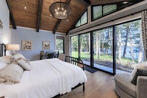 Ein Schlafzimmer mit einem großen Bett, einem Schreibtisch und Blick auf einen Wald durch große Fenster.