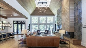Amplia sala de estar con un sofá grande, dos sillones, una mesa de centro y una chimenea. La habitación cuenta con suelo de madera, un techo alto con vigas a la vista y ventanas grandes que ofrecen vistas al exterior.