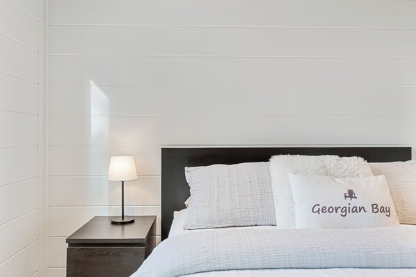 A neatly made bed with a white comforter and pillows, a bedside lamp, and a nightstand with a dark wood finish. - Play, dine and relax amongst the beauty of Georgian Bay and The Bruce Trail. (Lion's Head)