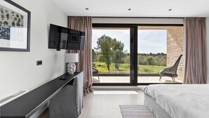 Ein modernes Schlafzimmer mit einem großen Bett, einem an der Wand montierten Fernseher, einem Schreibtisch mit Lampe und Blick auf einen Garten durch eine Schiebetür.