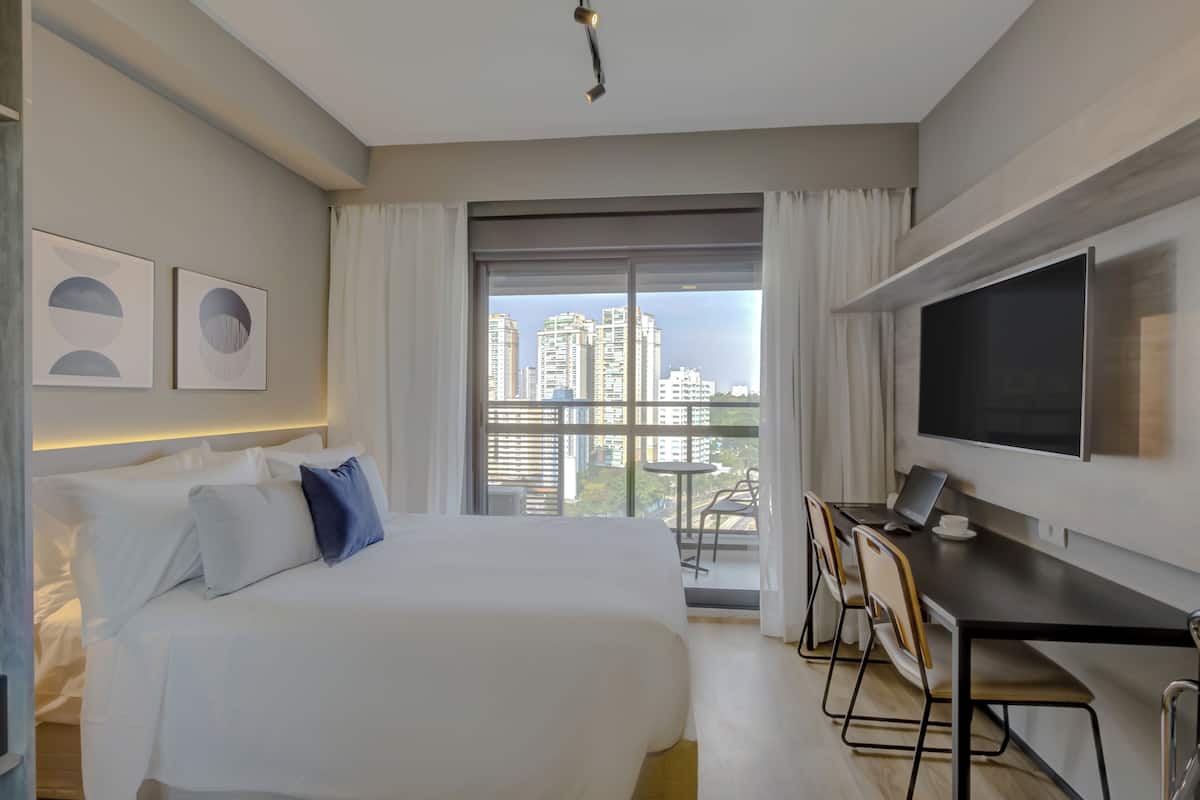 A modern hotel room with a large bed, a desk with a laptop, a flat-screen TV, and a balcony with a city view.