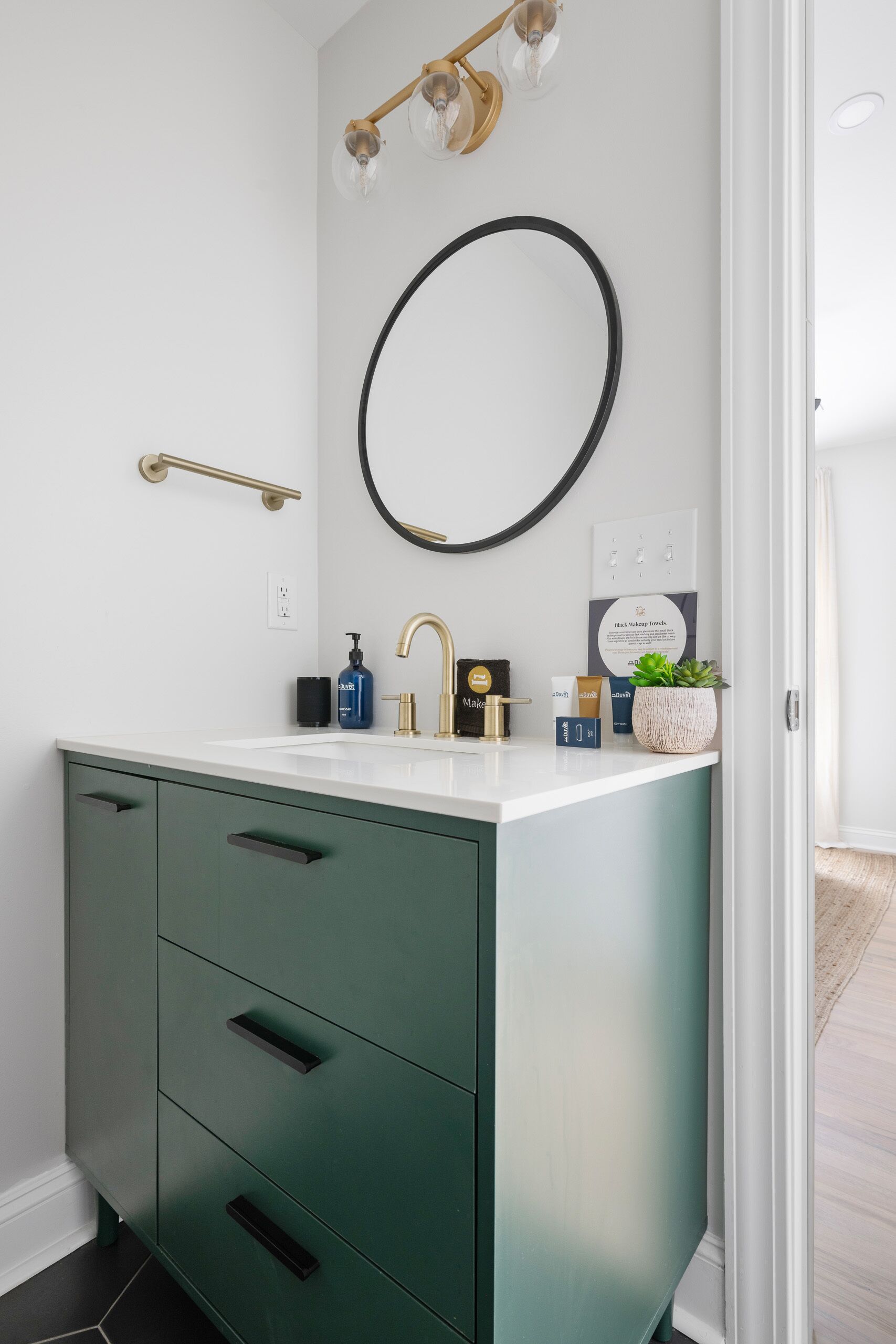 Een badkamer met een witte wastafel, een groen kastje en een ronde spiegel. Er staan toiletartikelen op het aanrecht en een plantje op de wastafel.