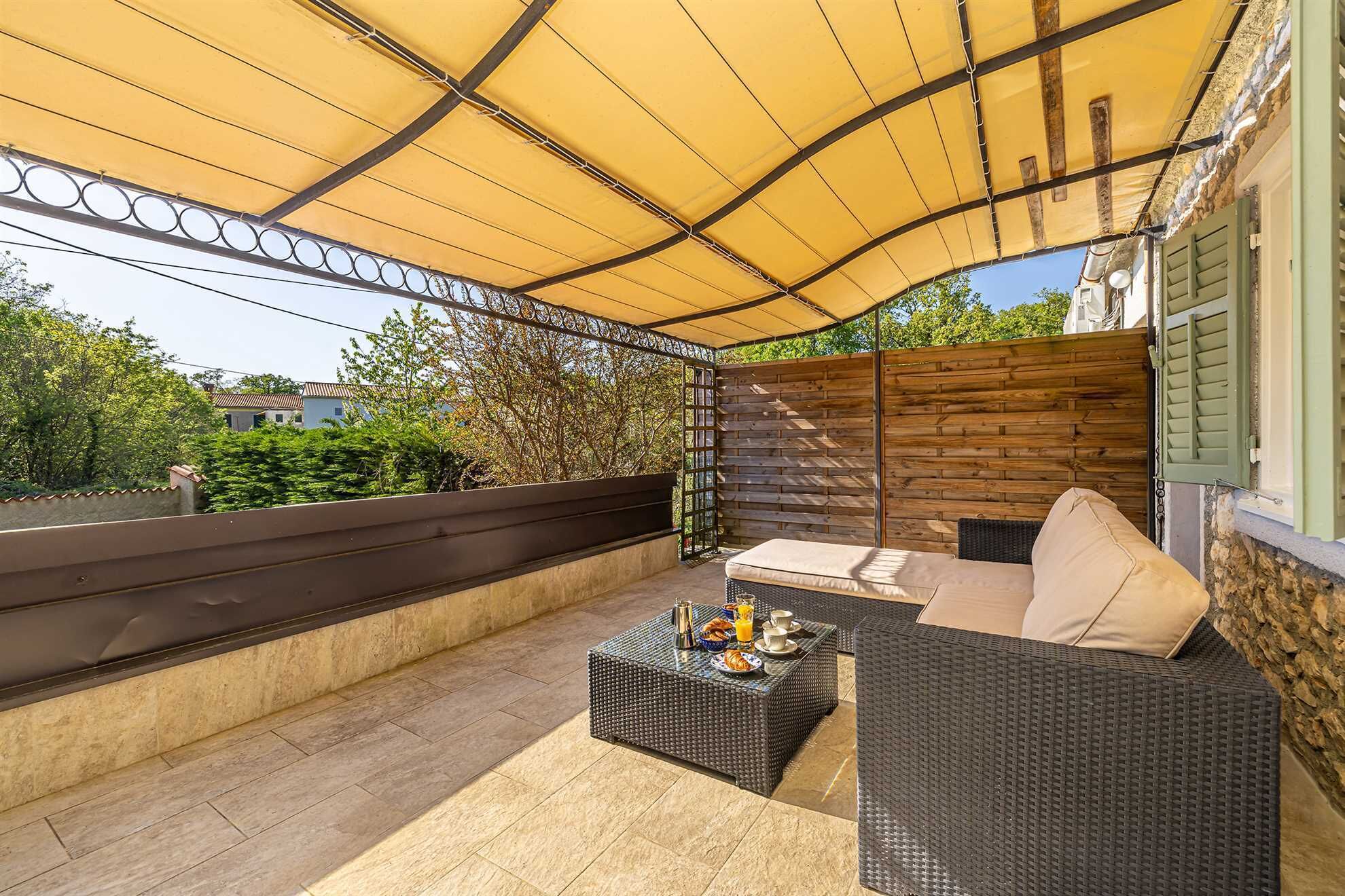 A covered outdoor seating area with wicker furniture, a table with breakfast items, and a view of greenery and buildings in the background.