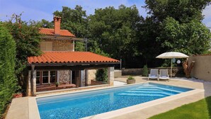 A swimming pool with a stone deck, a covered patio, and a lounge area with chairs and an umbrella.
