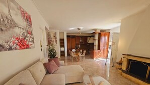 Ein Wohnzimmer mit einer Couch, einem Glastisch und einem Essbereich mit Stühlen und einem Tisch. Im Hintergrund befindet sich ein Kamin und eine Küche.