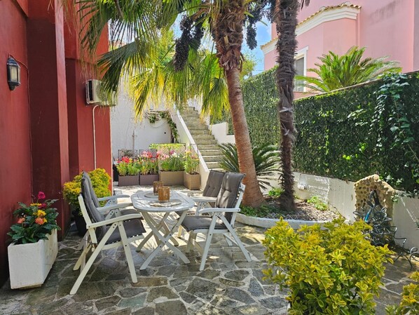 A patio with a stone floor, white chairs, and a table. There are potted plants and a staircase leading to an upper level.