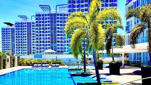 A swimming pool with lounge chairs, palm trees, and modern buildings in the background. - Luxury 1BR 55sqm 1 Pacific MactanNewtown (Lapu-Lapu)