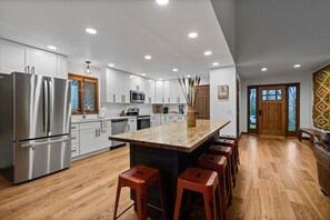 Une cuisine moderne avec des meubles blancs, un réfrigérateur en acier inoxydable, un îlot central en bois avec des tabourets, et une grande fenêtre avec des stores.
