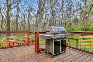 Ein tragbarer Grill auf einer Holzterrasse mit Blick auf Bäume und einem eingezäunten Bereich mit roten Stühlen.