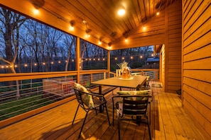 An outdoor wooden deck with a table and chairs, string lights, and a view of the surrounding trees.