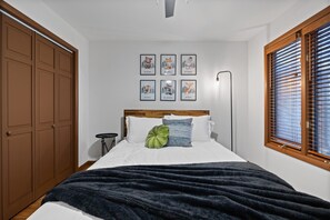 Ein Schlafzimmer mit einem Bett, einem grünen Kissen, einer dunklen Decke, einem hölzernen Kopfteil, einer Wand mit gerahmten Bildern, einem Fenster mit Jalousien und einem Kleiderschrank.