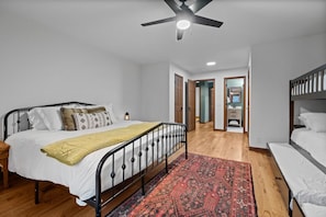 Un dormitorio con cama, mesita de noche, ventilador de techo, alfombra roja estampada y piso de madera.