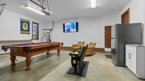 A pool table with a projector, a television, and a refrigerator in a room with white walls and a wooden door.