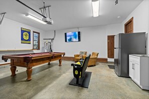 Une table de billard, un projecteur, un téléviseur et un réfrigérateur dans une pièce aux murs blancs et à la porte en bois.