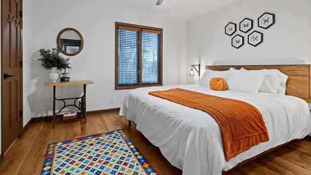 Ein Schlafzimmer mit Bett, Deckenventilator, Fenster mit Jalousien, Spiegel, kleinem Tisch mit Pflanze und farbenfrohem Teppich.
