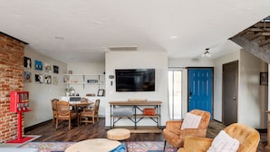 A living room with a brick wall, a dining area with a table and chairs, a large flat-screen TV mounted on the wall, and a blue door in the background. - Family Home with Private Hot Tub by Lake Hefner (Oklahoma City)