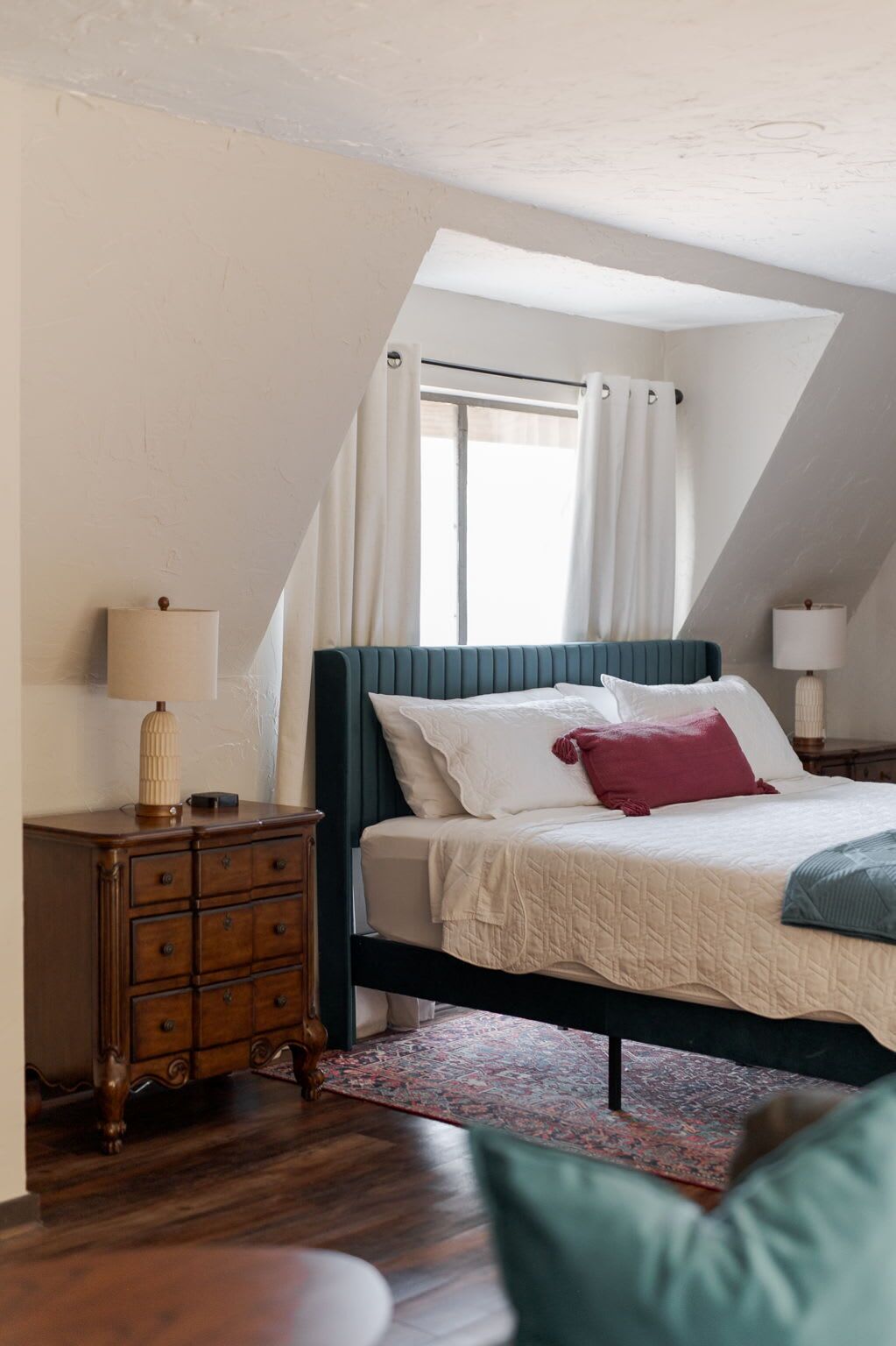 A bedroom with a bed, a dresser, a nightstand, a lamp, and a window with curtains.