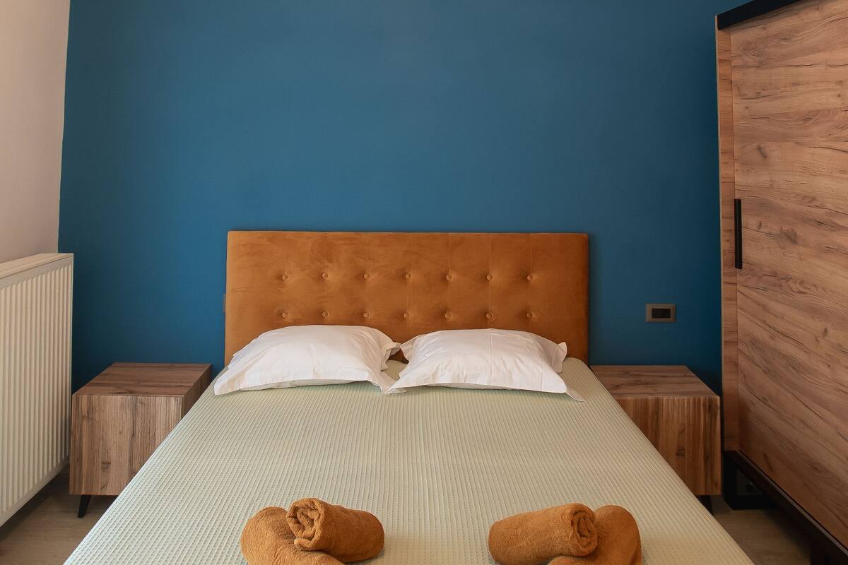 A neatly made bed with two rolled towels, a tufted headboard, and a blue wall.