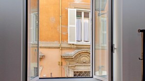 A view through a window with a white frame, looking out at a building with a yellow facade and a white window with shutters. - 4BNB - Lungaretta Jacuzzi Apartment (Rome)