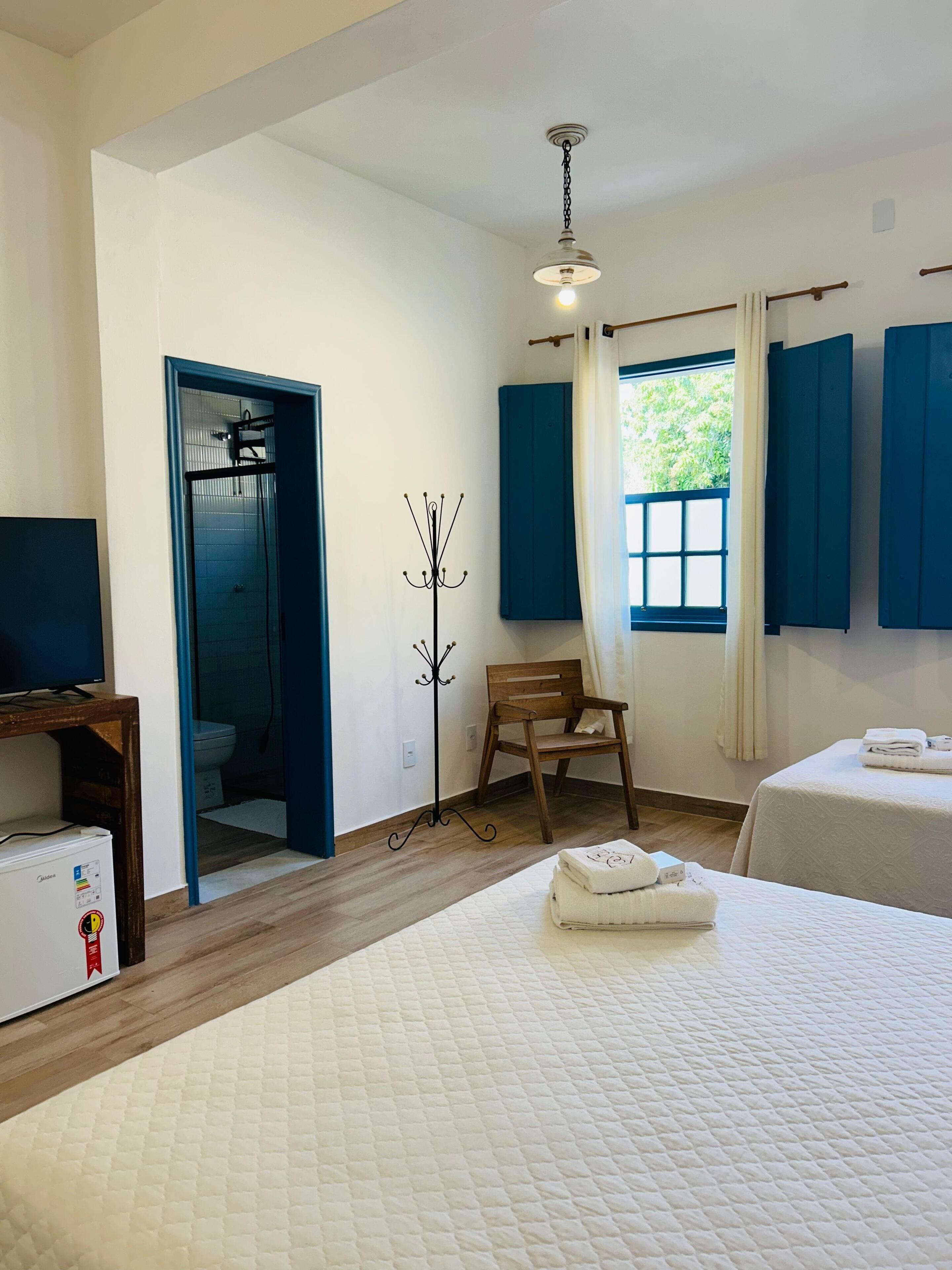 A hotel room with a bed, a chair, a coat rack, a window with curtains, and a ceiling fan.