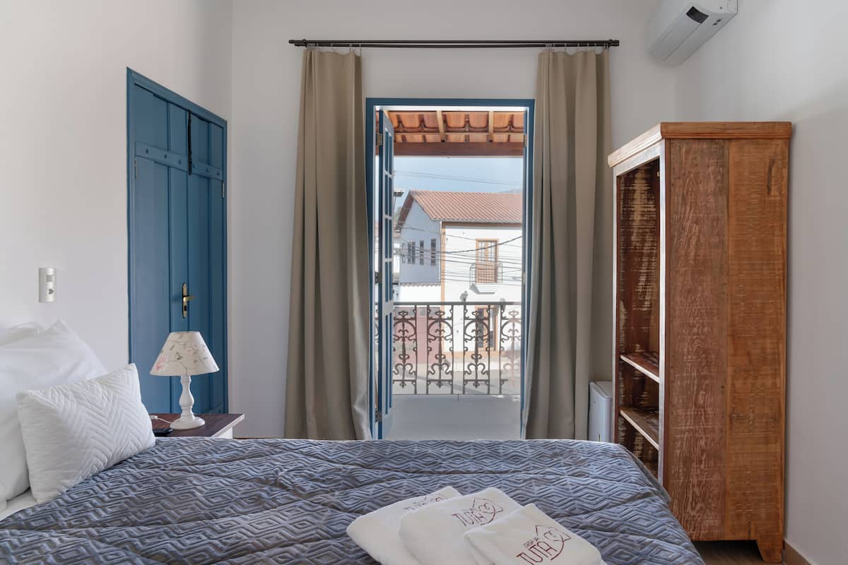 A bedroom with a bed, a nightstand, a lamp, a wardrobe, and a view of the outside.