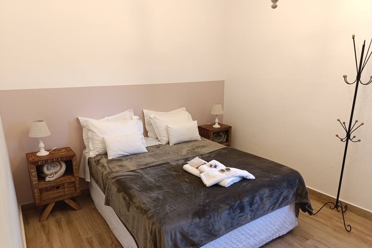A neatly made bed with a dark grey blanket, white pillows, and a folded towel on the bed. There are two bedside tables with lamps and a decorative item on one. The room has wooden flooring and a plain wall.
