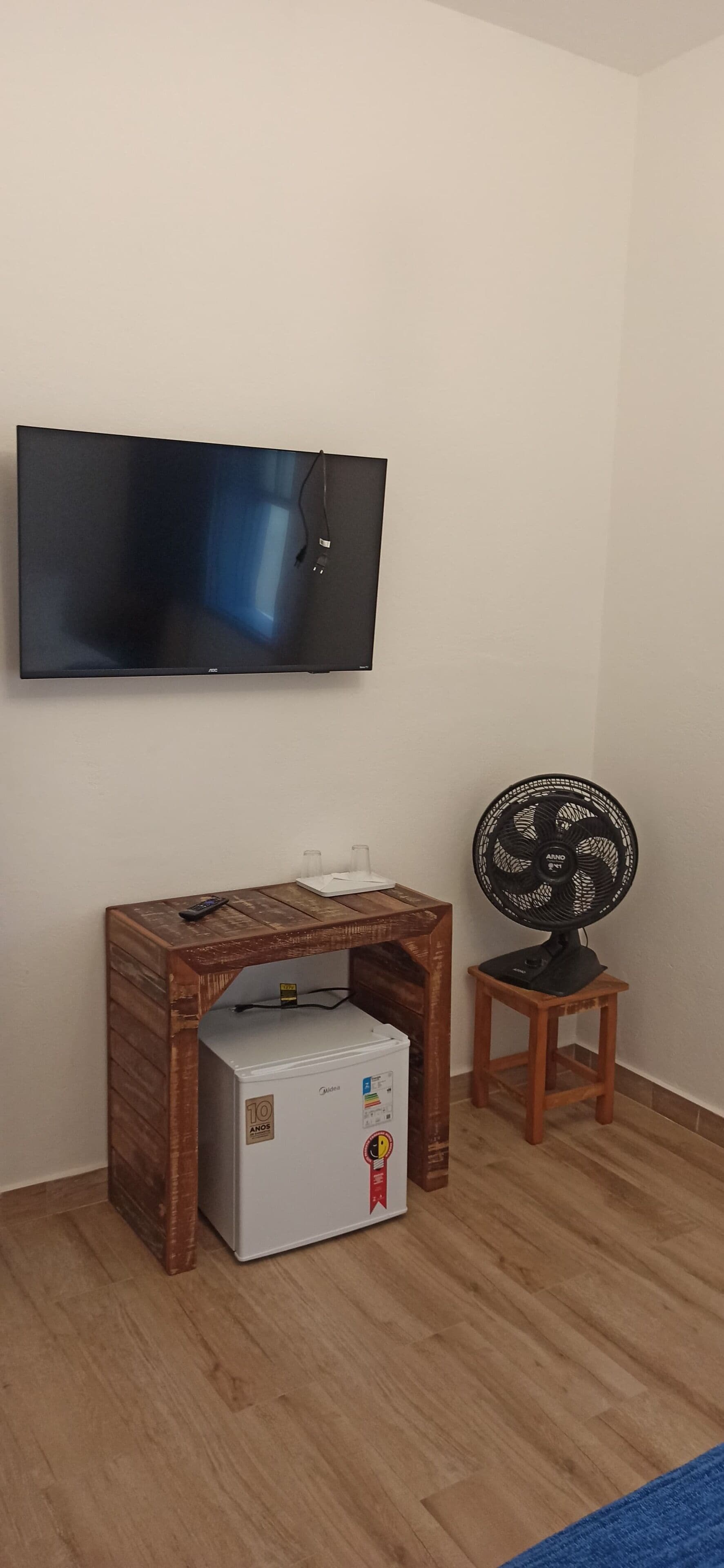 Een kamer met een wandgemonteerde flatscreen televisie, een houten tafel met een kleine koelkast en een staande ventilator.