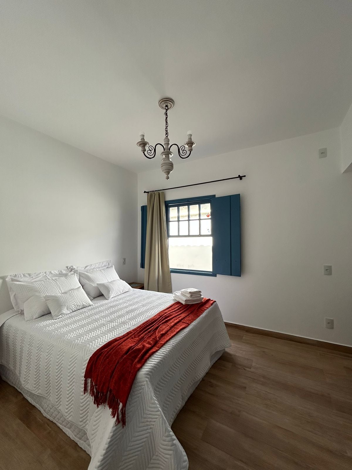 A bedroom with a bed, a window with blue shutters, a chandelier, and wooden flooring.