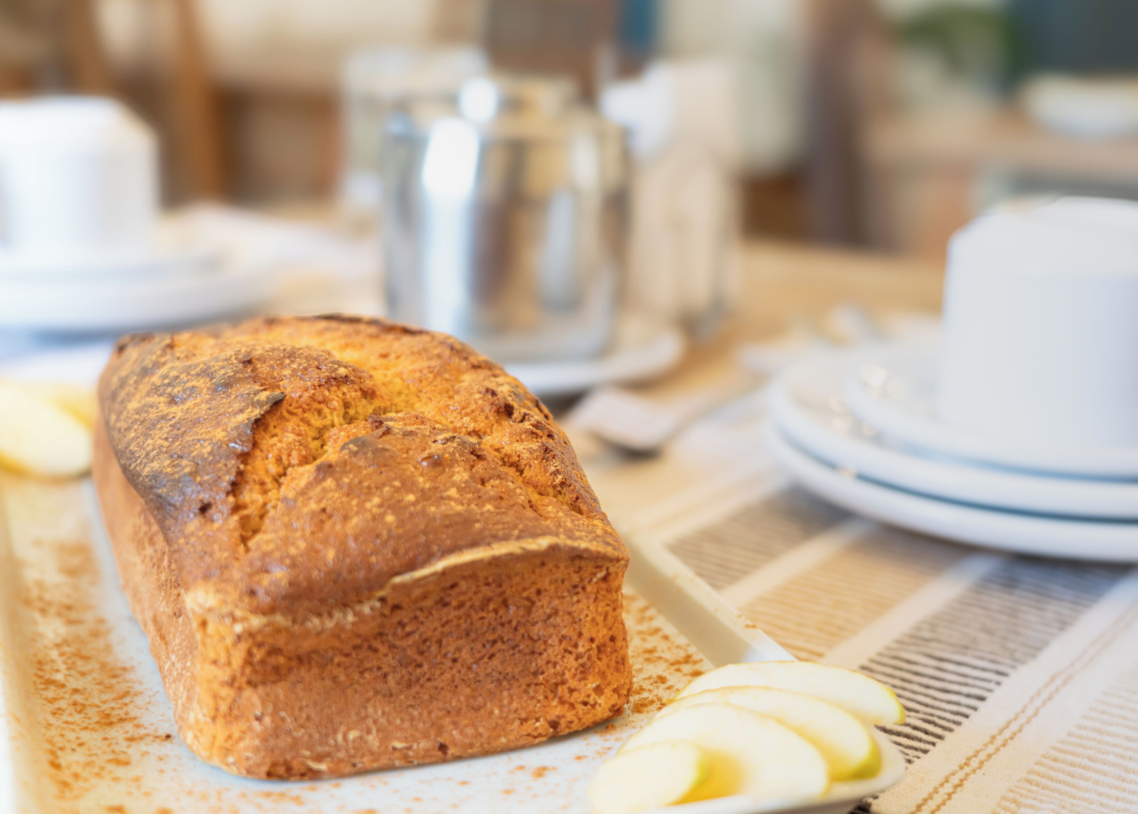 Ein Brot auf einem Tisch mit einer gestreiften Tischdecke, dazu Apfelscheiben und eine weiße Tasse.