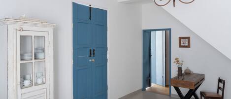 Una habitación con un armario blanco, una puerta azul, una mesa de madera con una silla y una araña.