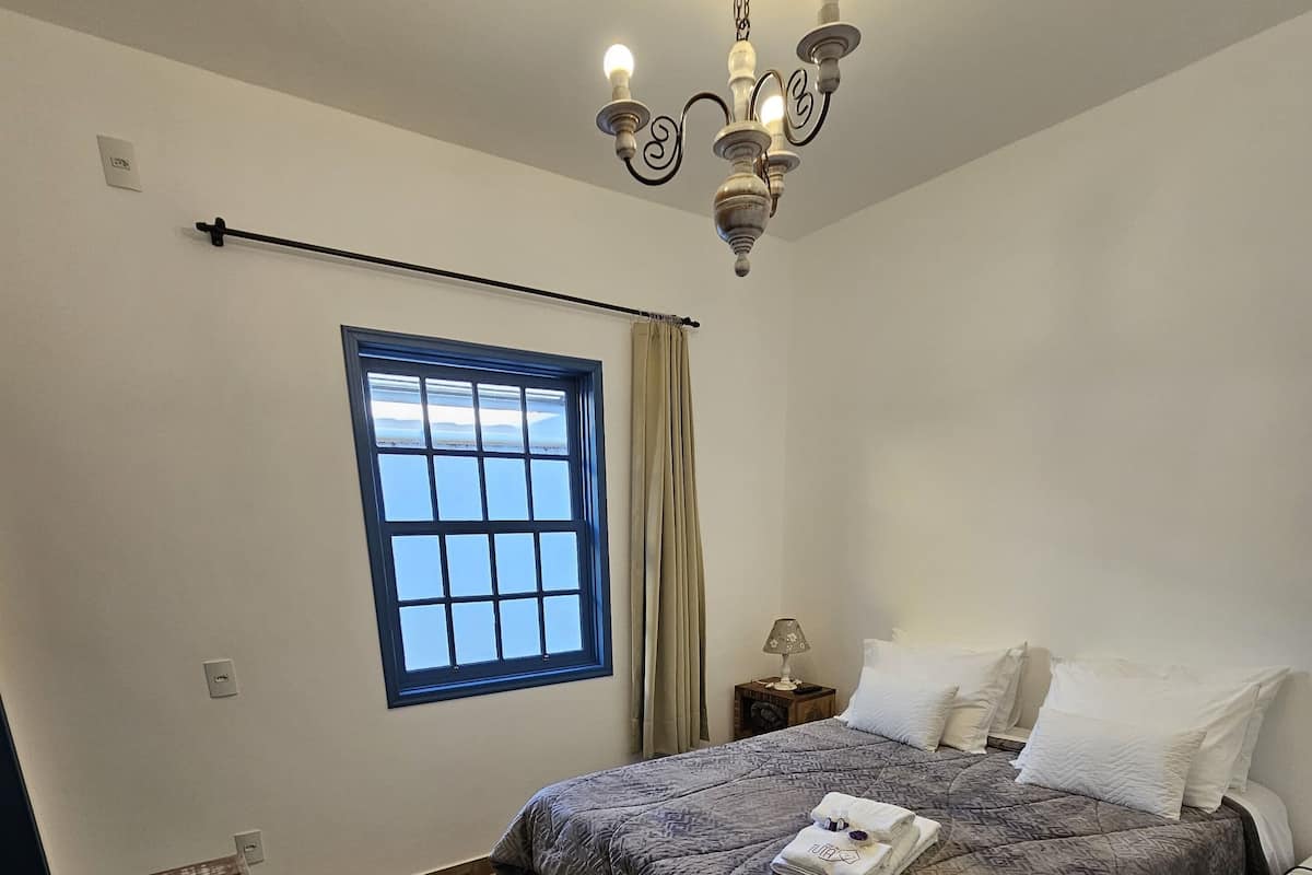 A bedroom with a large bed, a wooden dresser, a window with curtains, and a chandelier.